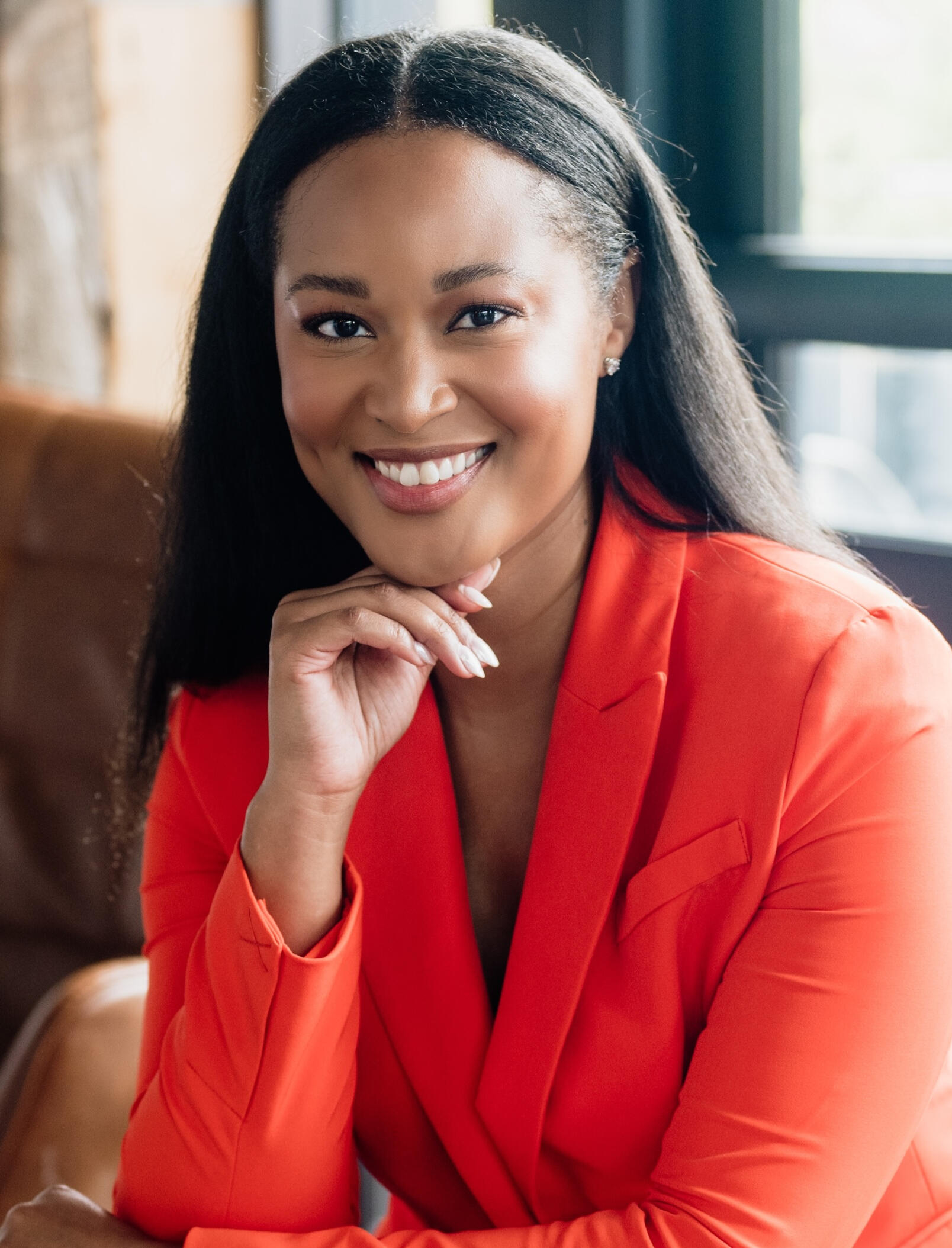 Tara Lassiter Headshot Tara Lassiter, seen smiling wearing a bright orange blazer, seated indoors near a window, resting her chin on her hand with a relaxed and confident expression.
